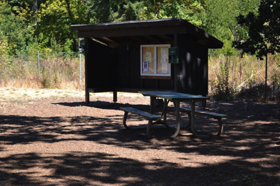 A covered kiosk, a dog waste bag container and picnic bench inside the off–leash pet area